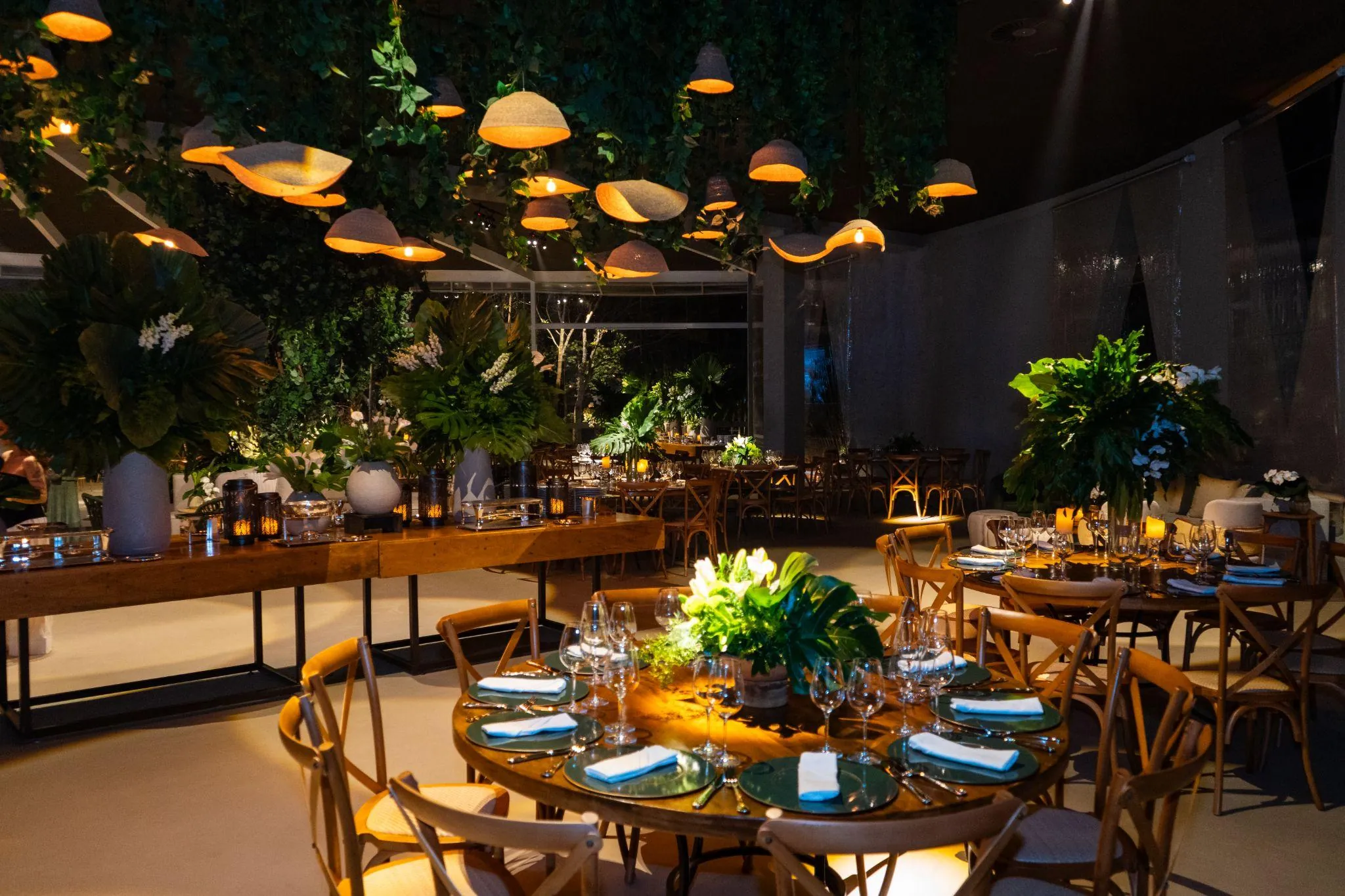 Salão de jantar com luminárias suspensas e mesas postas entre plantas